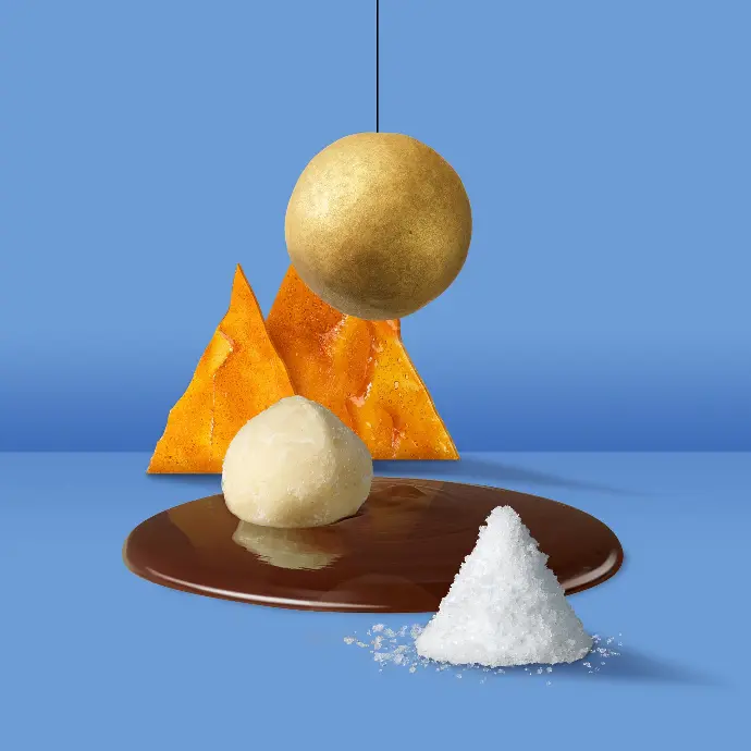 Maison Macolat golden chocolate enrobed macademia nut over a chocolate pool and some caramel triangles  on a blue background