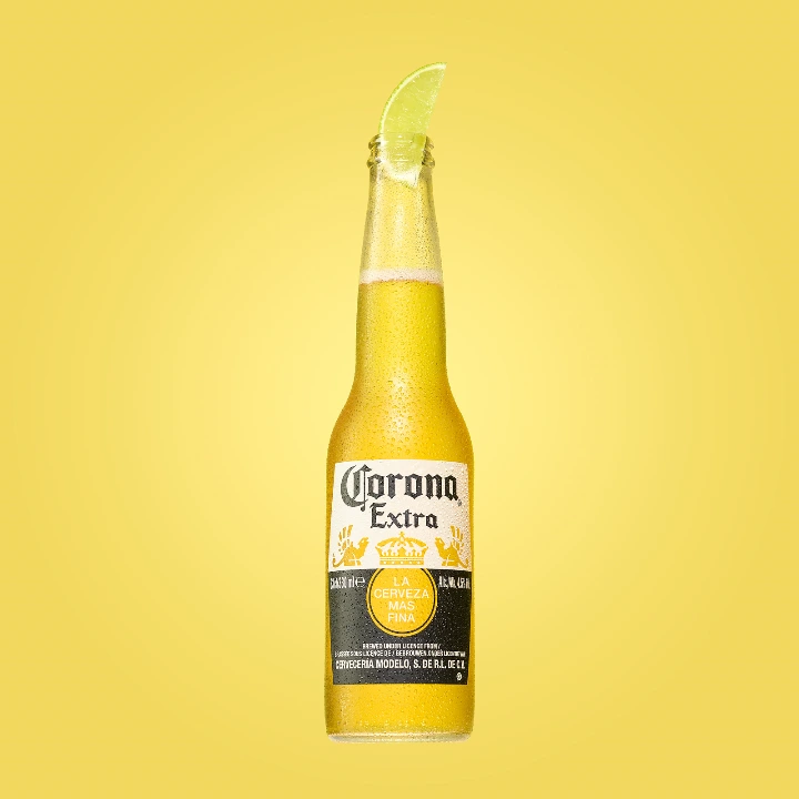 Corona Extra beer bottle with a lime wedge on the rim and real condensation droplets on the chilled glass, photographed against a warm yellow background. Drinks photography by Studio Ballena.