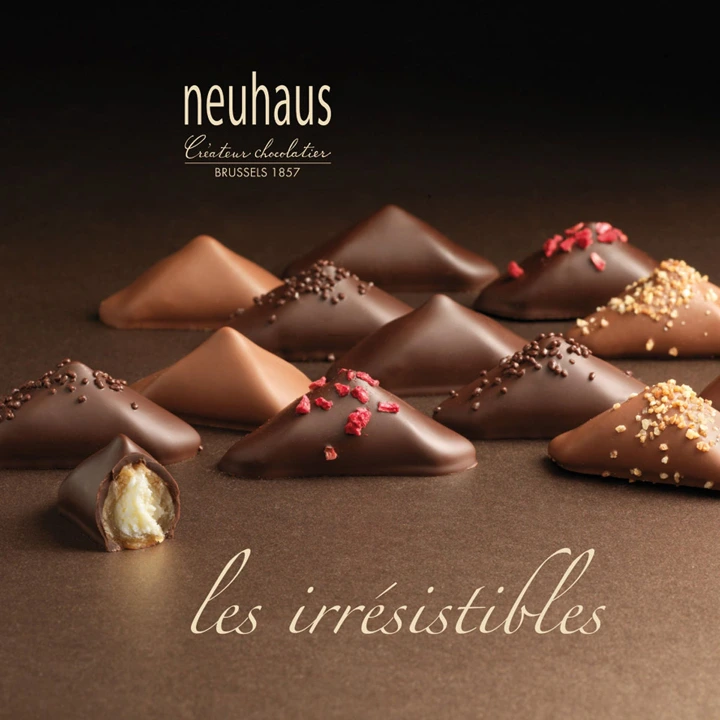 Neuhaus 'les irrésistibles' chocolate truffles — pyramid-shaped chocolates in dark and milk finishes, with sprinkles, chopped nuts and red flakes, on a dark surface. One truffle bitten in half showing the cream filling. Advertising photography by Studio Ballena for Neuhaus (Brussels, 1857).
